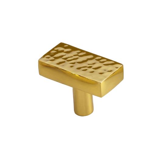 A polished gold cabinet knob with a rectangular top featuring a textured, hammered pattern, mounted on a cylindrical base. The knob is displayed on a white marble surface with gray veining.