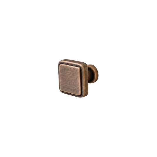 A square cabinet knob with a brushed metal finish in an antique bronze or gold tone, mounted on a plain off-white wall.